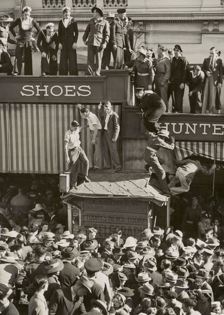 Civilians and servicemen celebrate in a city street, climbing the awnings of businesses below
